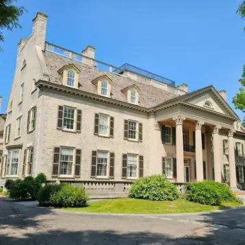 Large historic mansion, the George Eastman museum, with columns and many windows, framed by green trees under a bright blue sky.