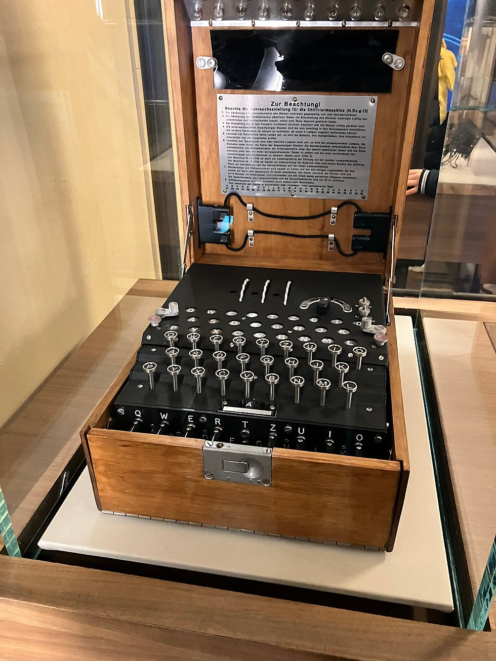 Wooden Enigma machine with open lid, showing keys and rotors. Black and silver details. German text plaque visible. Displayed in a glass case.