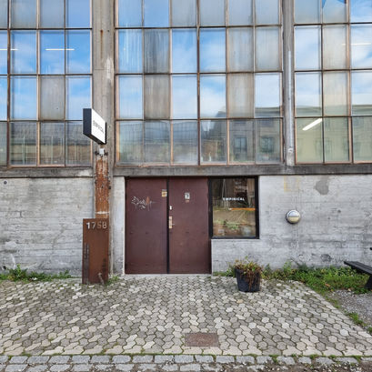 Industrial building facade with large glass windows and brown doors. Sign reads "Empirical." Paved walkway in front, mood is neutral. The home of the Empirical Spirits brand and visitor experience, and tour and tasting room.