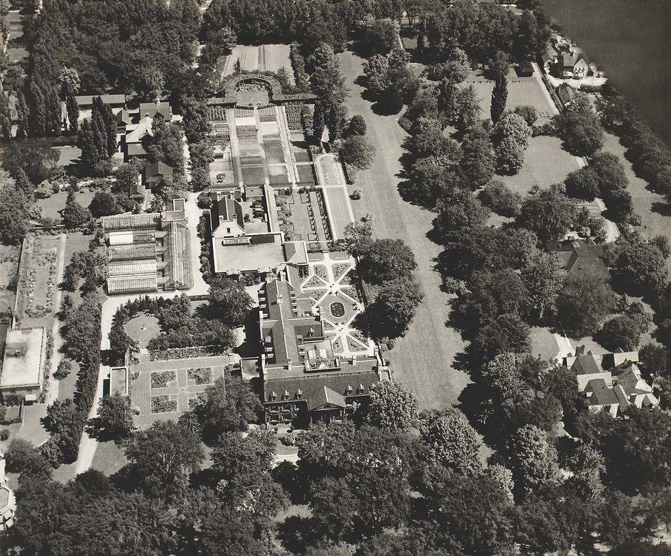 Aerial view of a grand estate with manicured gardens, greenhouses, and tree-lined grounds beside a river.