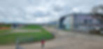 A person in a red coat walks near the modern museum building for the Silverstone Museum, under a cloudy sky. Green grass and a Union Jack mural are visible.