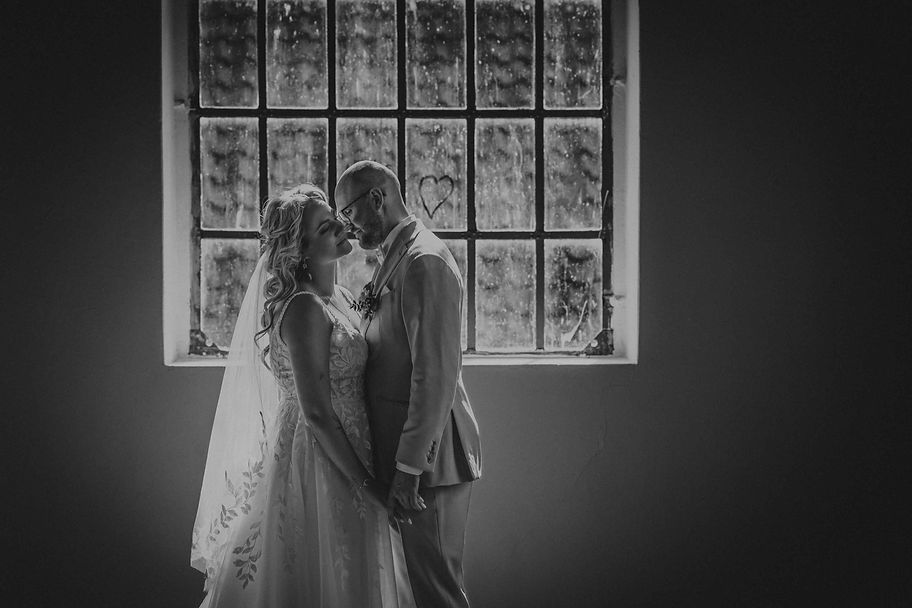 Schwarzweiß‑Hochzeitsfoto eines Brautpaares vor einem alten Fenster; die Braut im Spitzenkleid und Schleier, der Bräutigam im hellen Anzug, beide eng beieinander in einem intimen Moment – fotografiert von Tino Hochzeitsfotograf aus Braunschweig.