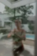 Ein atmosphärisches Foto einer Person durch eine Glasscheibe, die einen Topf mit Pflanzen hält. Im Hintergrund ist ein moderner Innenhof mit Pool zu sehen, umgeben von Grünpflanzen und einer weißen Mauer