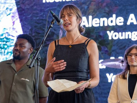 Three people stand before a colorful background. Sammy Weaver in the center performs into a microphone, holding papers. Text reads "Alende Amisi Young Poet."