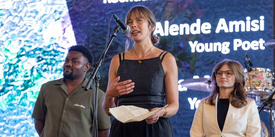Three people stand before a colorful background. Sammy Weaver in the center performs into a microphone, holding papers. Text reads "Alende Amisi Young Poet."