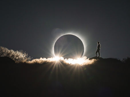 A total solar eclipse just above a ridge with a sunstar bursting through the vegetation and a person standing in silhouette
