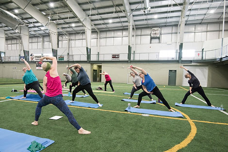 An indoor group essentrics class on green turf. The instructors back is to the camera, and the participants are facing the camera. They are all in a side lunge with one arm reaching overhead.