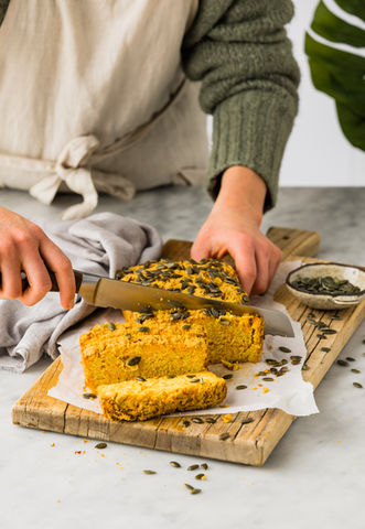Person cutting pumpkin bread on a wooden board atop a marble tabletop, wearing a linen apron, with a monstera leaf peeking in the background.
