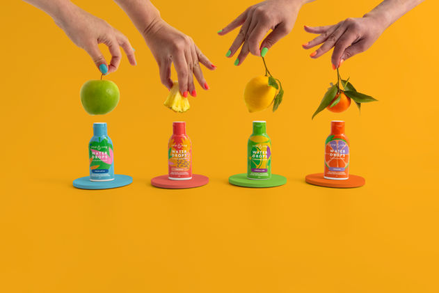 Four hands holding fresh fruits above the flavoured water drops bottles on a colour-block yellow background.