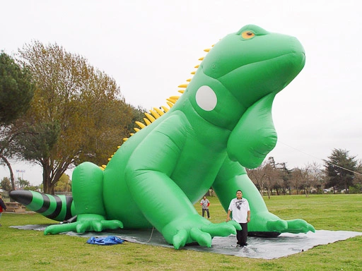 Side view of a large inflatable iguana set up in a park with a person standing beside it for size reference.