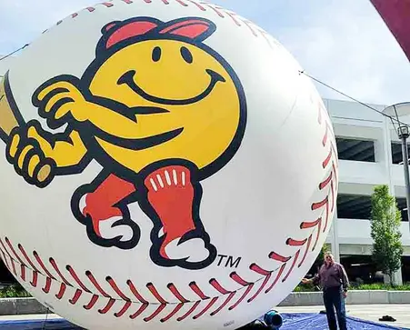 Large inflatable baseball with a smiley-faced batter mascot at an outdoor sports event.