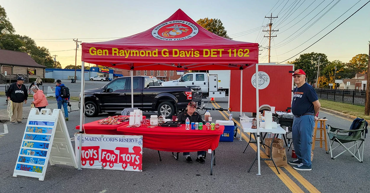 Red heavy duty canopy tent with Marine Corps branding and Toys for Tots table set up on a street for a charity event.