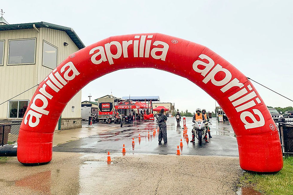 Red Aprilia inflatable finish line arch at a rainy motorcycle track with cones, staff, and riders approaching beneath it.