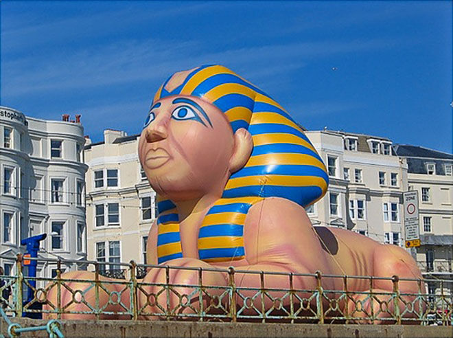 Large Egyptian sphinx inflatable landmark outdoors with a bright blue sky and multi-story buildings behind it.