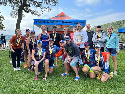Athletic team posing in front of a branded orange and blue pop up canopy at an outdoor race event near a lake.