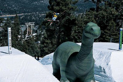Giant inflatable brontosaurus dinosaur displayed in a snowy mountain environment, with a long neck silhouette visible above the snow