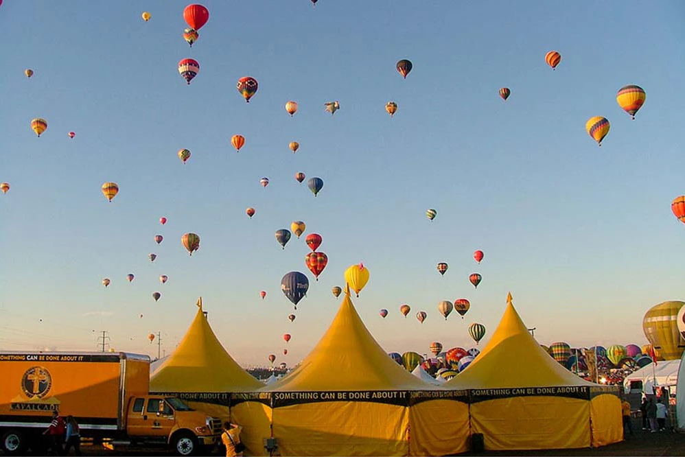 Series of yellow high peak event tents used for branding at a hot air balloon festival with dozens of balloons in flight.