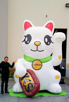 White inflatable lucky cat statue holding a gold coin, displayed at an event with a person standing next to it.