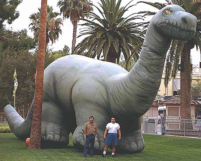 Giant inflatable brontosaurus dinosaur installed on grass with palm trees for scale at an outdoor event setup