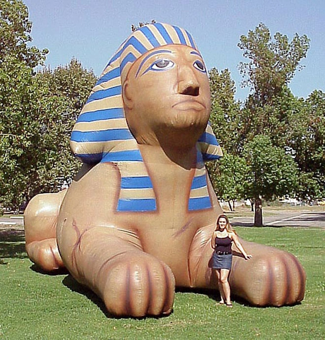 Giant inflatable sphinx on a grassy field with a person standing beside it for scale.