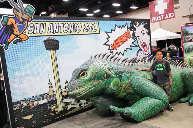 Giant inflatable iguana set up indoors beside a San Antonio Zoo backdrop with a person standing near it for scale.