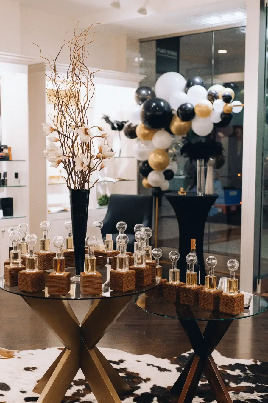 Elegant perfume boutique grand opening display with fragrance bottles on glass tables, black white and gold balloon decor, and modern luxury interior.