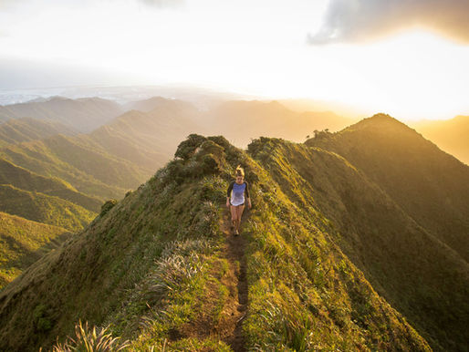 A person hiking on a narrow mountain ridge at sunset, surrounded by lush green hills and a golden sky, creating a serene, adventurous mood.