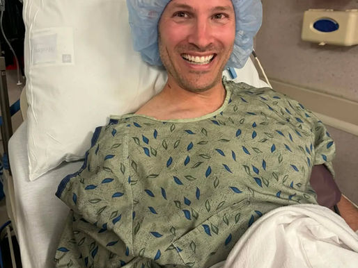 A man is lying down in a hospital bed wearing a hospital gown and surgical cap awaiting surgery.