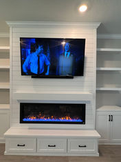 White custom-built entertainment center with shiplap detailing, TV mount, electric fireplace, and floating shelves, surrounded by storage cabinets, all beneath an intricate crown molding