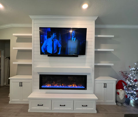 White custom-built entertainment center with shiplap detailing, TV mount, electric fireplace, and floating shelves, surrounded by storage cabinets, all beneath an intricate crown molding
