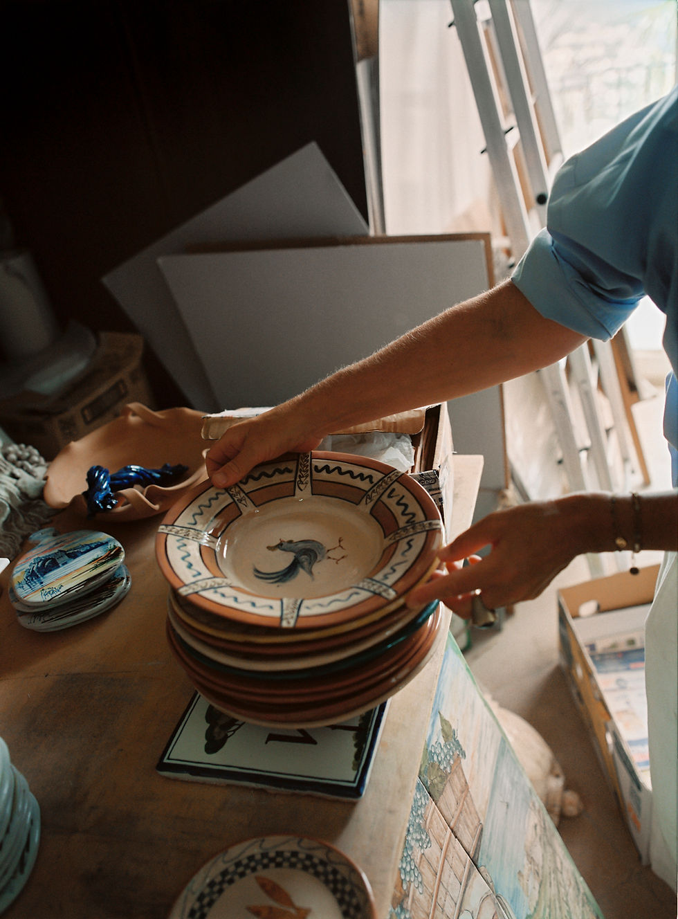 Person in blue shirt handles ceramic plates with rooster design in a cluttered room. Light filters through a window, ladder in background.