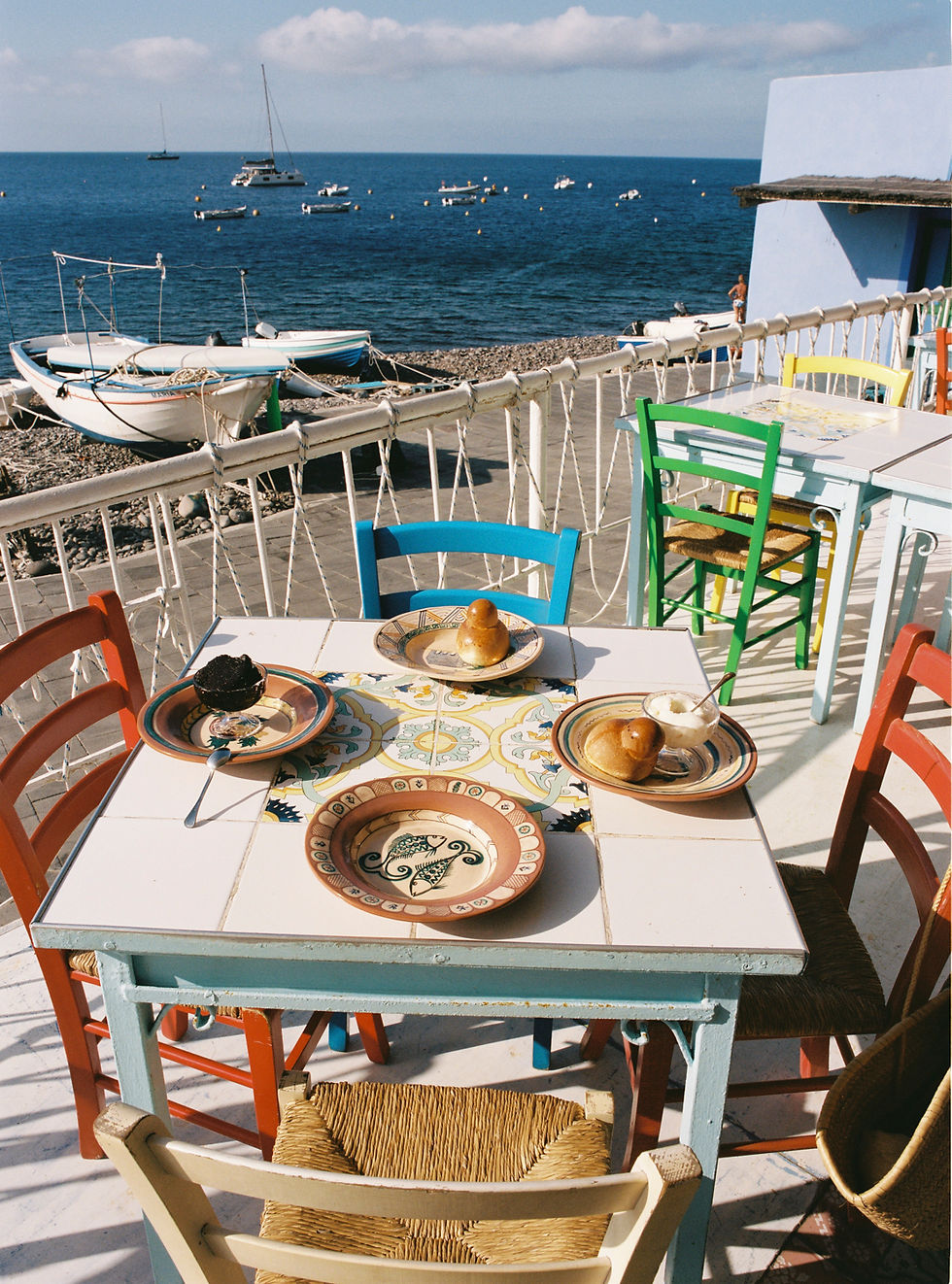 Colorful chairs surround a table with desserts on plates, set on a seaside terrace with docked boats and a blue ocean in the background.