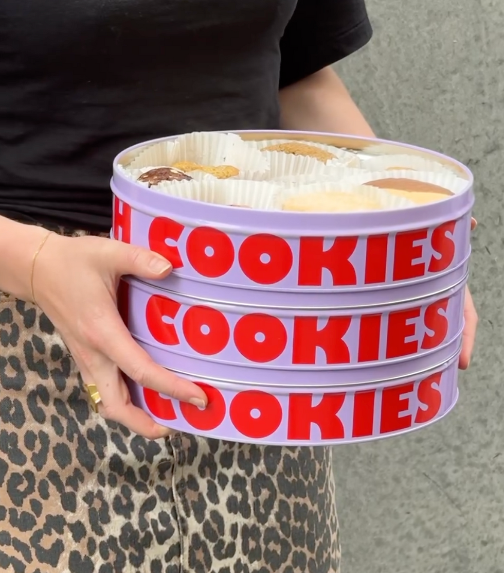 Person holds three purple tins labeled "COOKIES" in red, wearing a black shirt and leopard print skirt. Gray wall in the background. From Butterboy Manly.