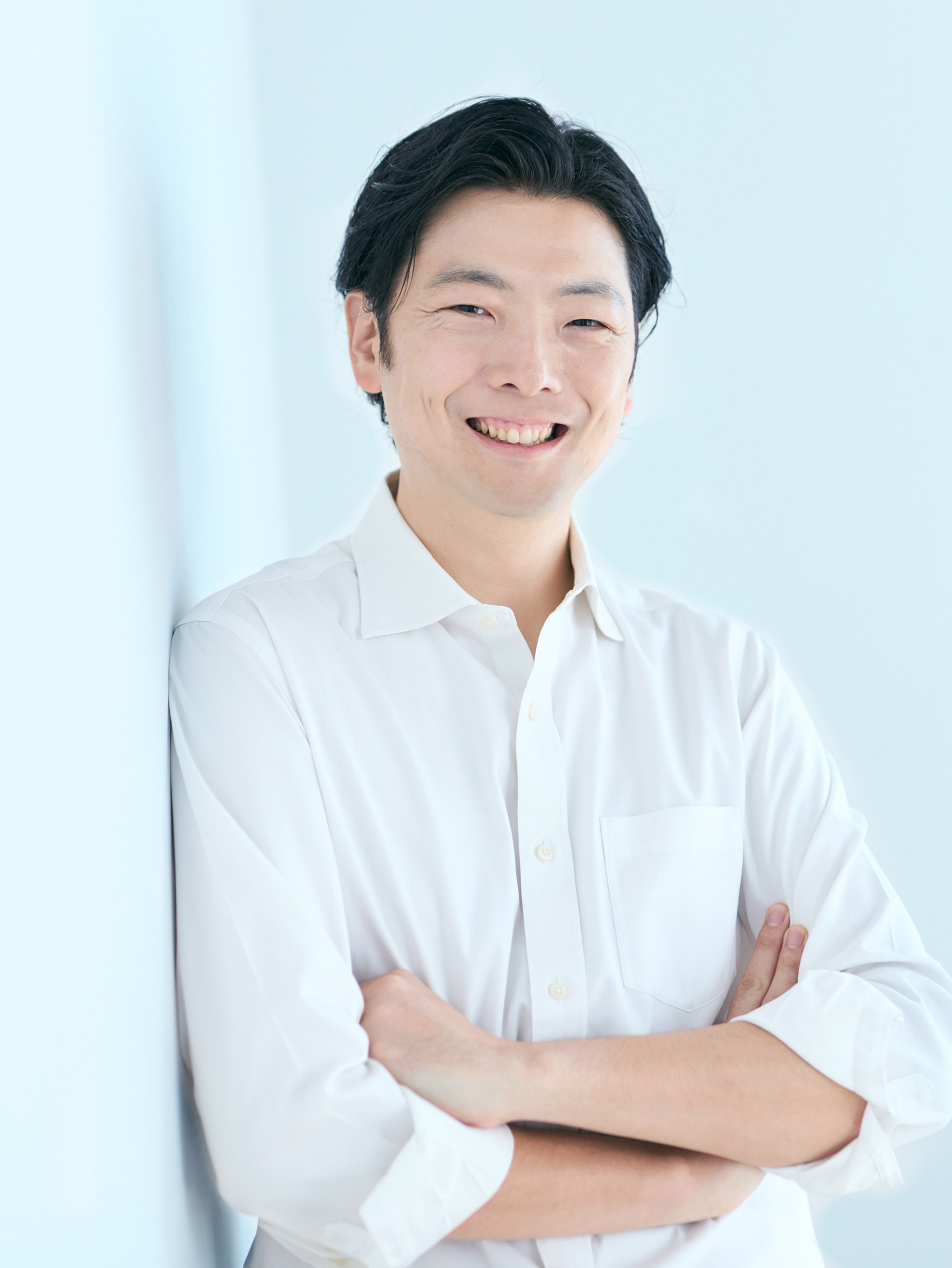 白いシャツを着た男性が淡い水色の壁際で腕を組み、笑顔でこちらを見る。明るく清潔感のある雰囲気。