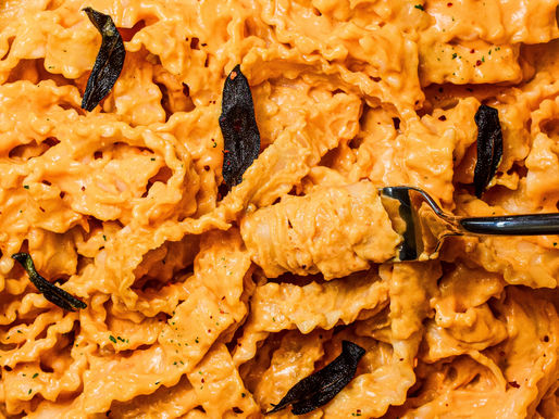 Creamy orange-hued pasta with sage leaves in a pan. A fork twirls the pasta, sprinkled with herbs and chili flakes, on a wooden table.