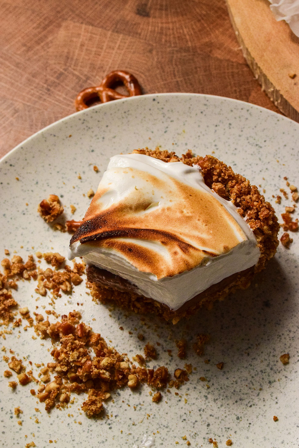 Toasted meringue dessert slice on a speckled plate with crumbs, beside a pretzel on a wooden table.