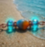 A close-up of a copper bracelet with glowing blue and orange beads resting on wet sand at the edge of the ocean.