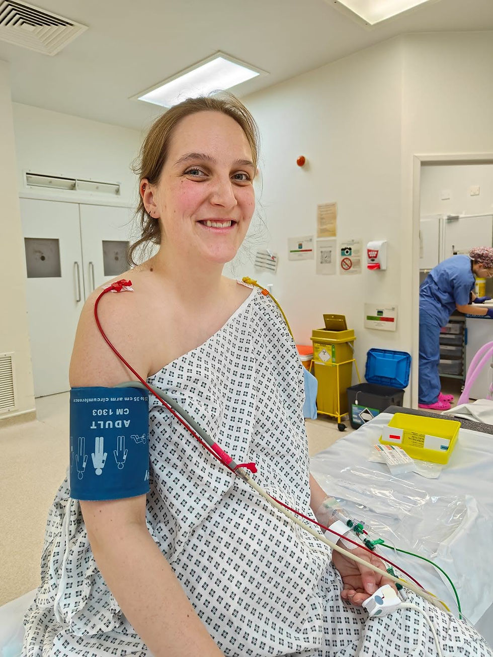 Smiling woman in a hospital gown, seated with medical wires attached, in a clinical room. Background shows medical equipment and a nurse.