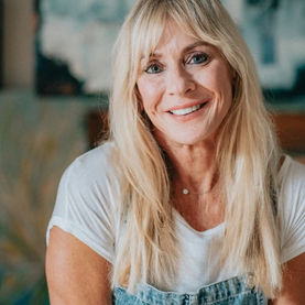 Blonde artist Kim Gibbs poses for her photo casually sitting in her art studio with an abstract painting in the background wearing her painting overalls 