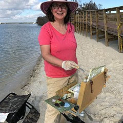 Artist Mary Hubley smiling while painting on the beach wearing a big sunhat and a bright pink shirt
