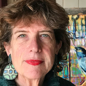 Artist Louanne LaRoche smiling for a very close up picture. She's wearing bright red lipstick and a folksy- style green and black earring. Shes posing in front of one of her colorful paintings and it looks like a painted bluebird is sitting on her shoulder