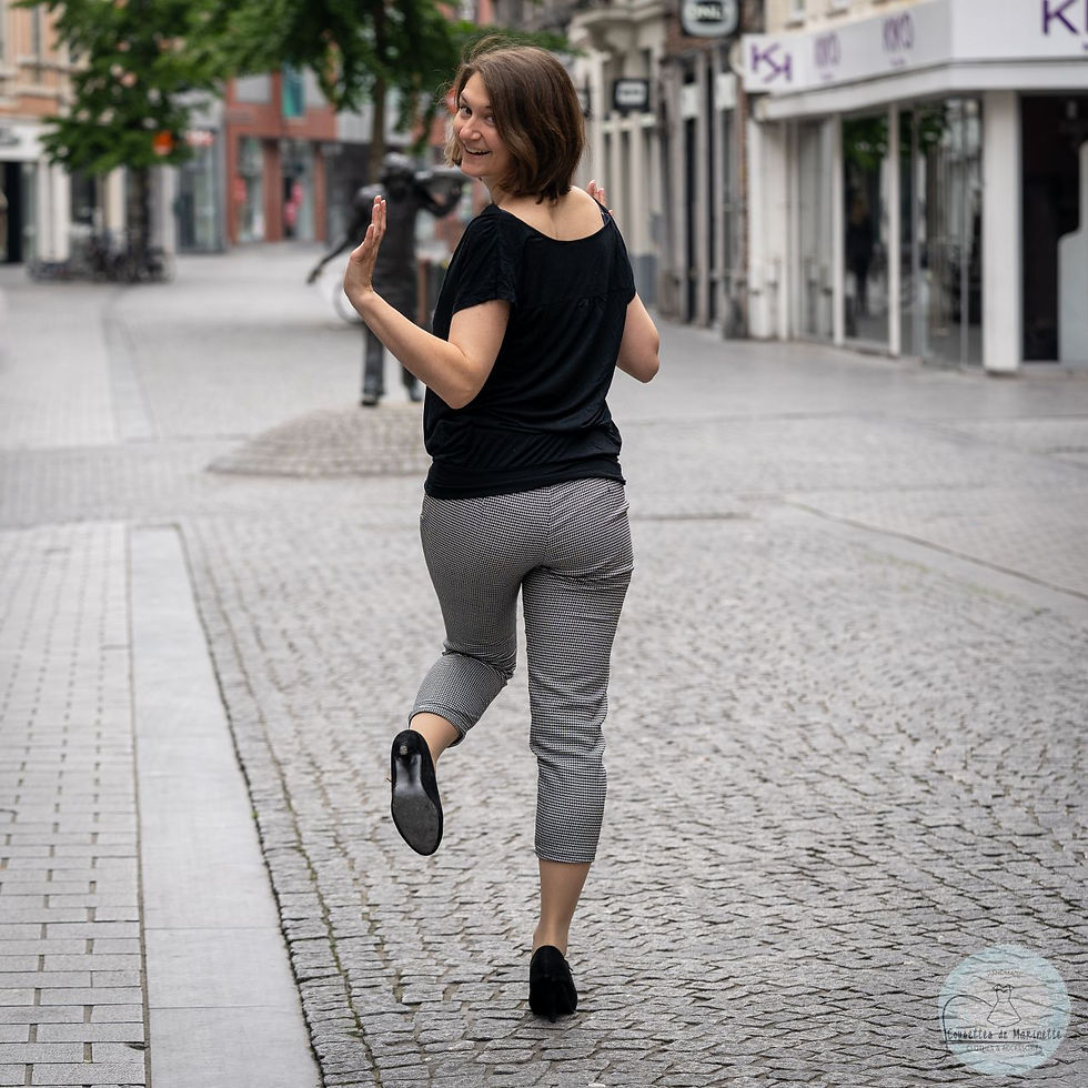 A woman with dark hair is dancing in the street, her back to the camera. She is wearing a black tee-shirt, black heels, and black and white calf-length trousers.