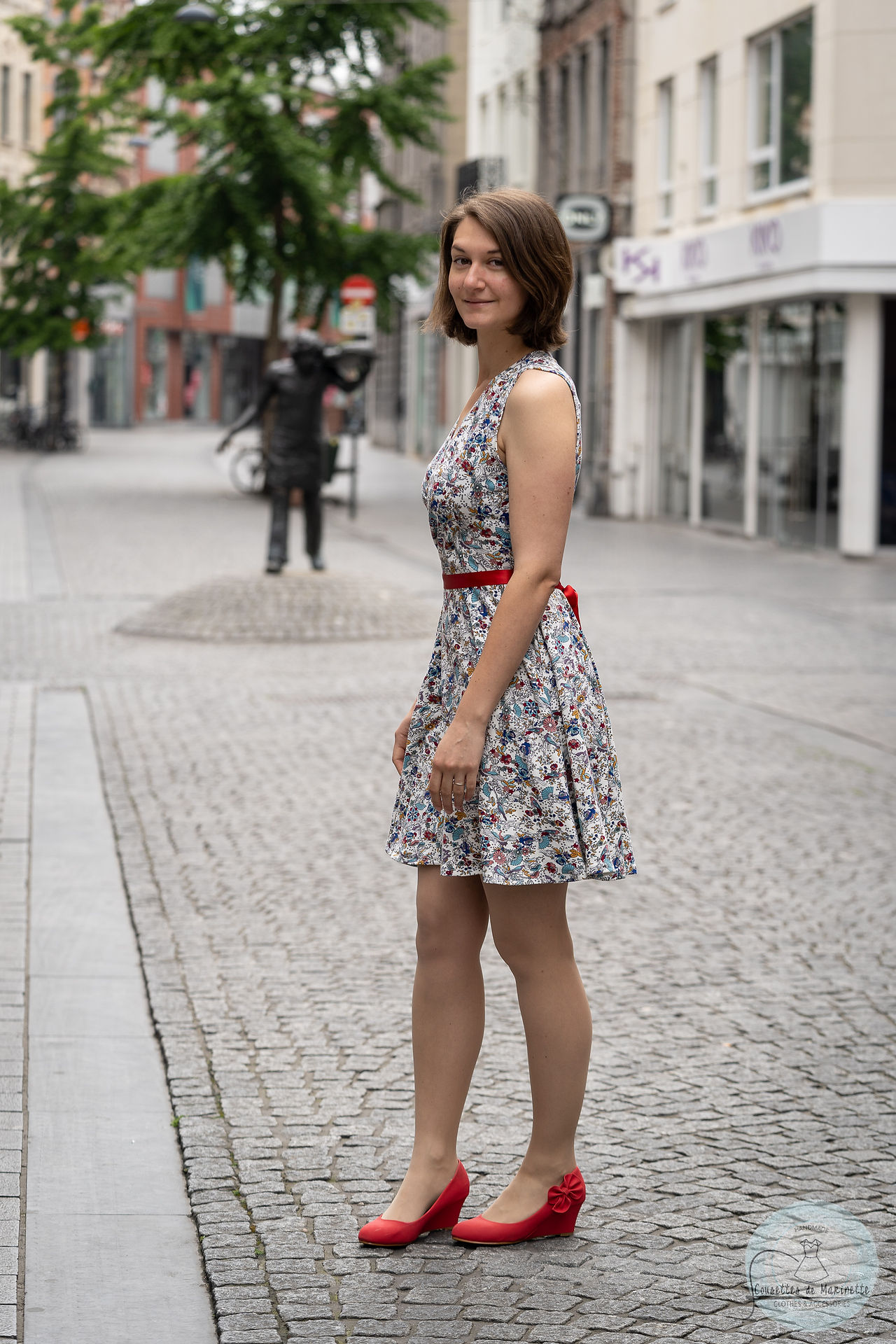 A woman with dark hair standing sideways in the street. She is wearing a white dress with lots of colourful characters on it, and a red belt