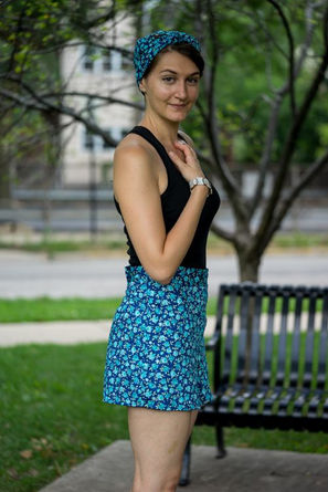 Une femme portant un débardeur noir, un short bleu à fleurs, des lunettes de soleil et un foulard dans les cheveux pose dans un jardin au soleil.