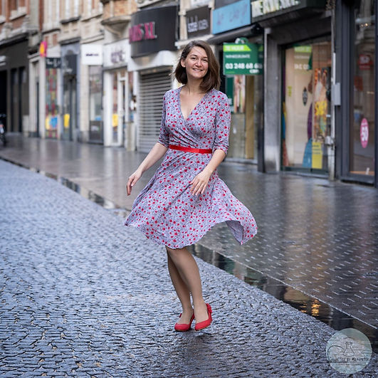 A woman is twirling in the street, while smiling at the camera. She is wearing a grey and red dress, with a red belt and red shoes.