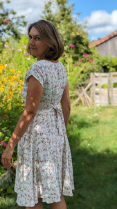 A woman with brown hair is wearing a white dress with small green and pink flowers. She is in a sunny garden surrounded by flowers, is turning her back to the camera and is looking above her shoulder