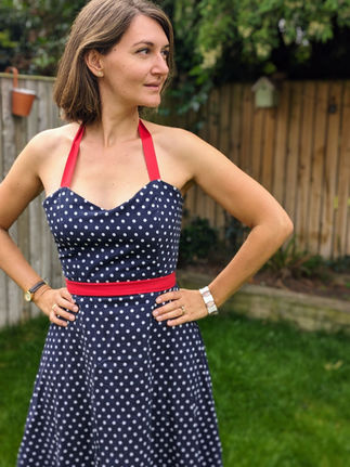 Une femme aux cheveux bruns mi-longs pose dans un jardin, devant une cloture en bois. Elle porte une robe bleue à pois blanc, avec une bretelle et une ceinture rouges. Elle se tient face à la caméra, les mains sur les hanches, et regarde sur le coté. dant l'objectif.
