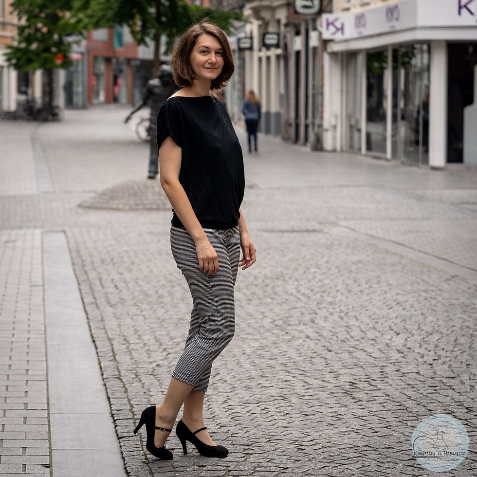 A woman with dark hair is standing in the street, sideways to the camera. She is wearing a black tee-shirt, black heels, and black and white calf-lenght trousers.