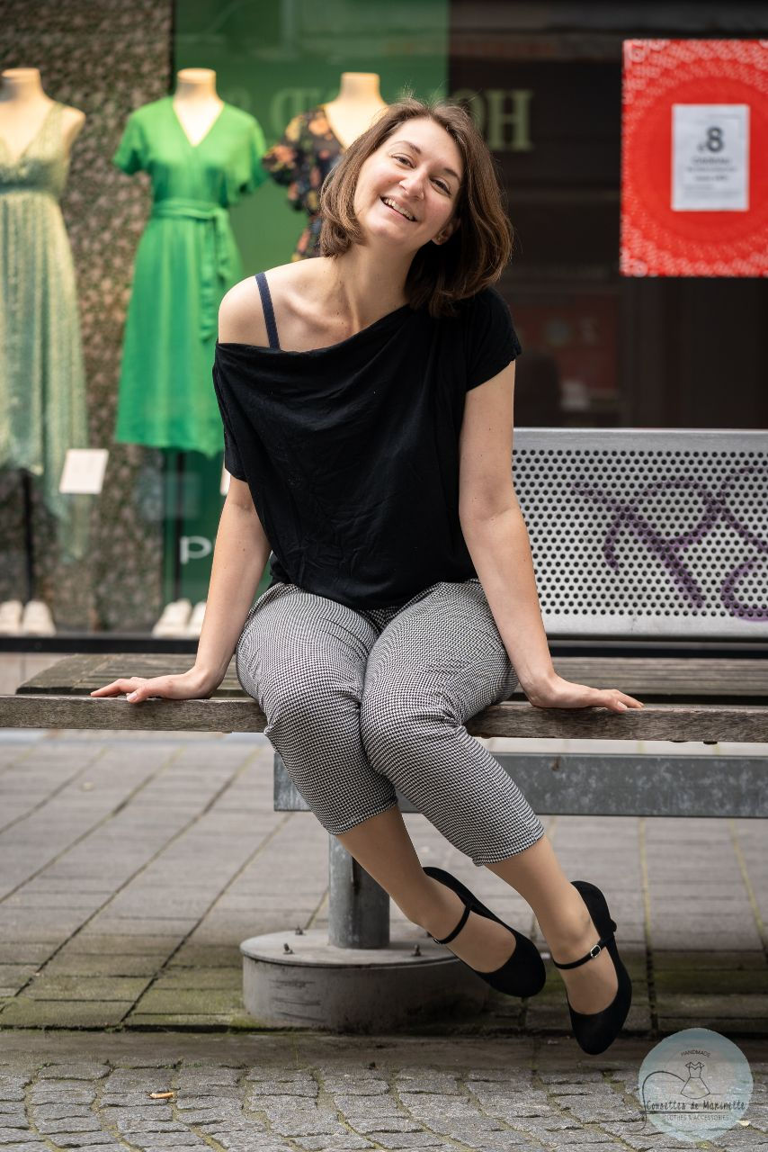A woman with brown hair is weraing a black tee-shirt and black and white trousers. She is sat on a bench, swinging her legs around