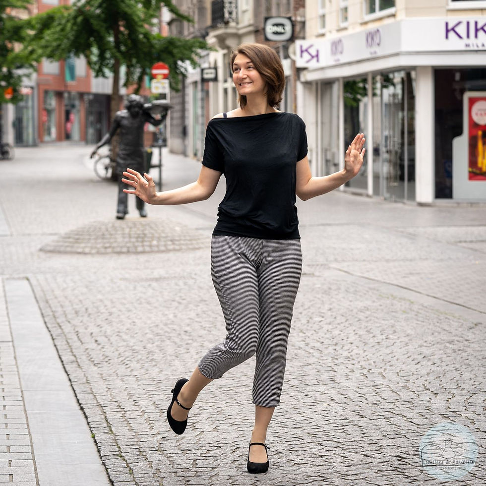 A woman with dark hair is standing in the street. She is wearing a black tee-shirt, black heels, and black and white calf-length trousers.